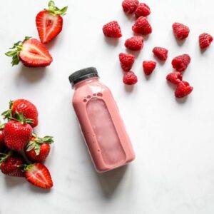 Top view of fresh strawberry and raspberry smoothie in a bottle with scattered fruits on white background.